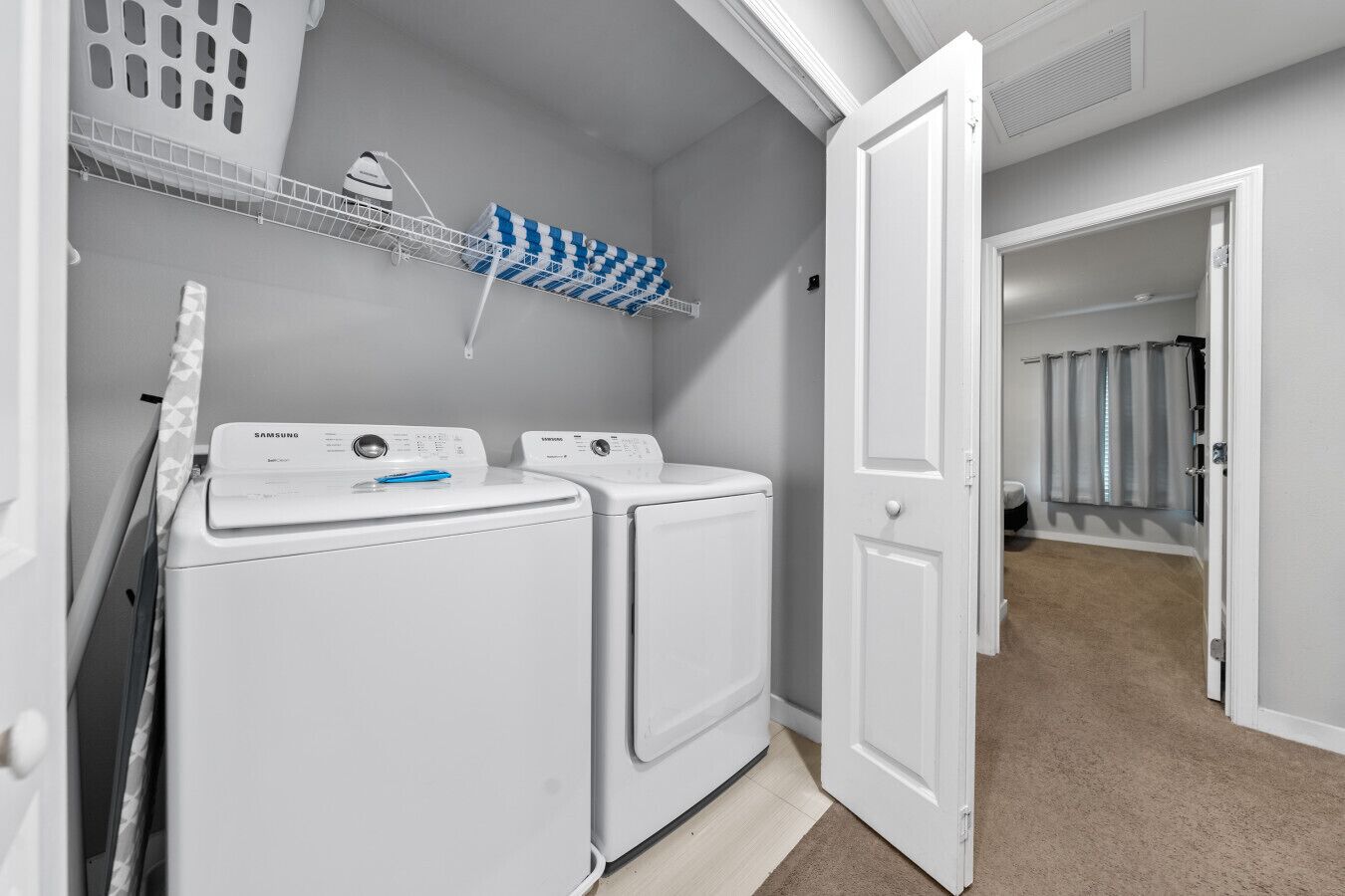 A laundry room with two white front-loading washers and dryers, a drying rack with blue and white towels, and a door leading to another room.