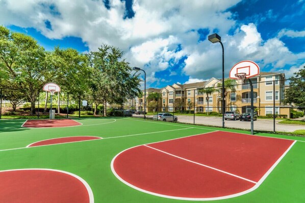 An outdoor basketball court with a green and red surface, surrounded by trees and a residential building in the background.