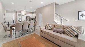 Een moderne woonkamer met een beige bank, een eethoek met een tafel en stoelen, en een keuken op de achtergrond.