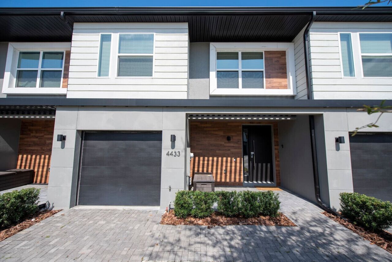 A modern two-story townhouse with a grey garage door, a wooden door entrance, and a number '4433' on the wall.