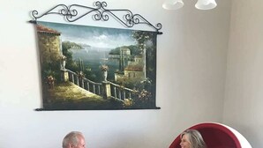 Two people seated in red chairs facing each other, with a painting of a coastal scene above them.