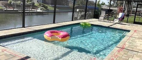 Une piscine avec un flotteur gonflable rose et jaune, entourée d’une zone abritée d’où l’on peut admirer un plan d’eau et des bâtiments en arrière-plan.