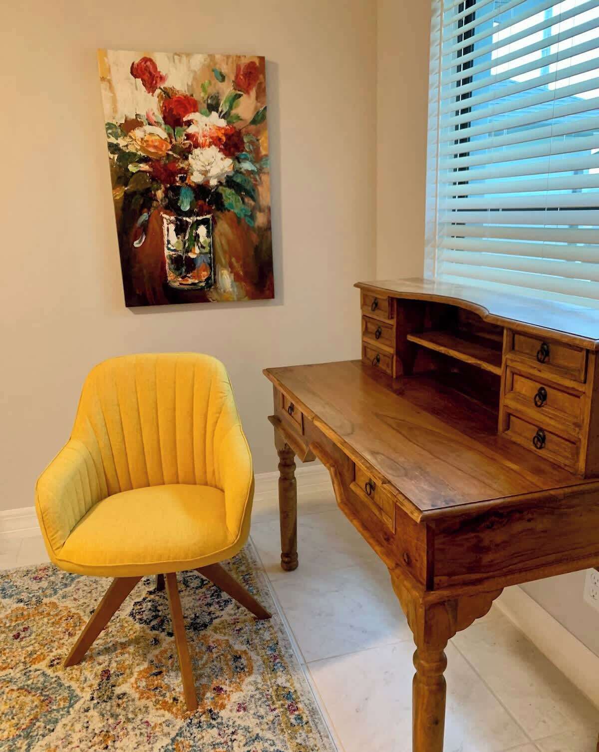 Una habitación con un sillón amarillo, un escritorio de madera con cajones, un cuadro de flores y una ventana con persianas.