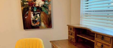 A room with a yellow armchair, a wooden desk with drawers, a painting of flowers, and a window with blinds.