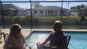 Two people seated at a table by a poolside, with a view of houses and a body of water in the background.