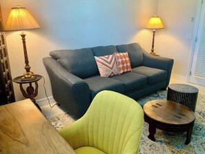 Ein Wohnzimmer mit einer Couch, einem grünen Sessel, zwei Lampen und einem kleinen runden Tisch.