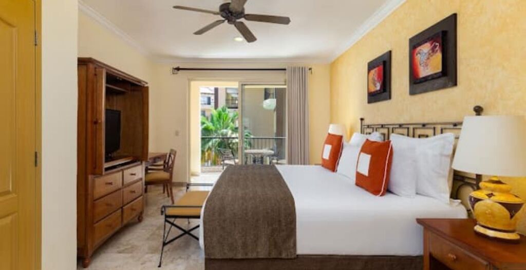 Une chambre d’hôtel équipée d’un lit, d’un bureau, d’une chaise, d’une télévision, d’un ventilateur de plafond et d’un balcon offrant une vue sur la verdure.