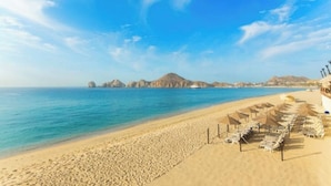 A beach with clear blue waters, a sandy shore, and rows of sun loungers with umbrellas. - Spectacular 2BD Beachfront Condo in Villa del Arco
 (Cabo San Lucas)