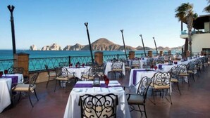 An outdoor dining area with tables set up for guests, overlooking a coastal view with mountains in the distance. - Spectacular 2BD Beachfront Condo in Villa del Arco
 (Cabo San Lucas)
