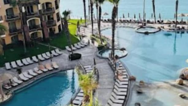 A resort with a pool area, palm trees, and a view of the ocean. - Spectacular 2BD Beachfront Condo in Villa del Arco
 (Cabo San Lucas)