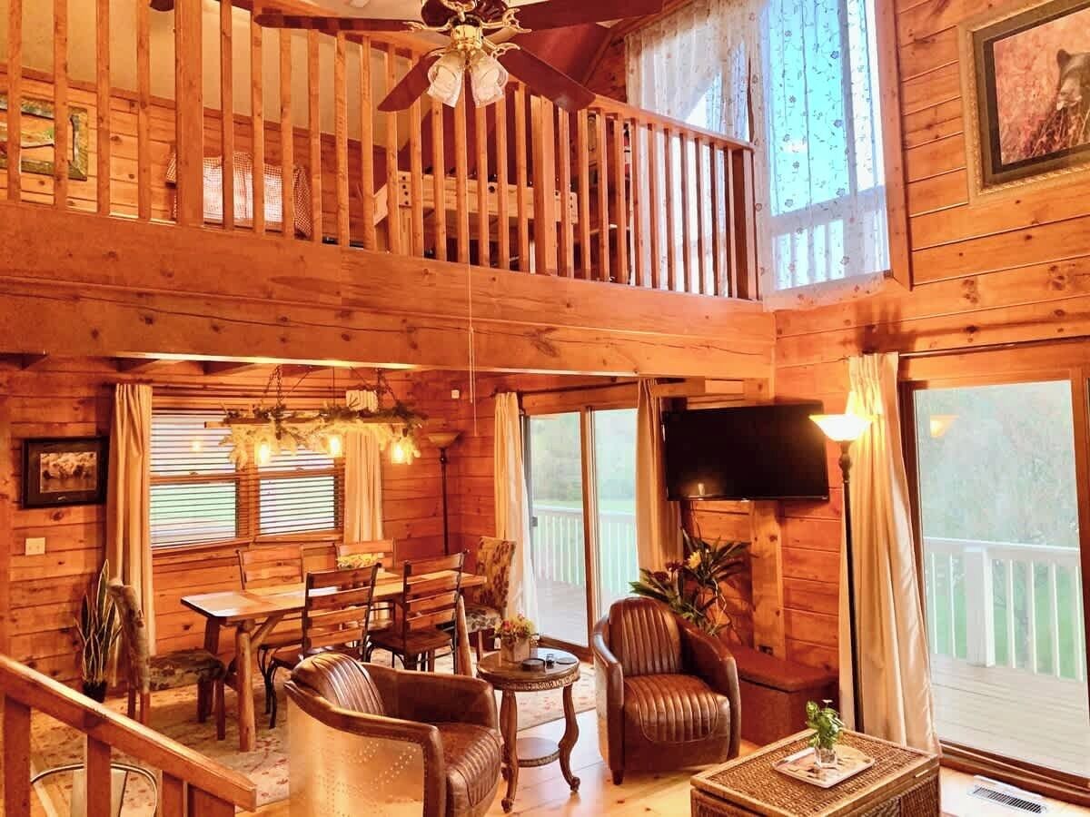 A wooden cabin interior with a dining area, a living room with a sofa and armchairs, a balcony, and a television.