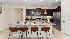 Een moderne keuken met een centraal eiland, roestvrijstalen apparatuur, hangende lampen en een witte deur.