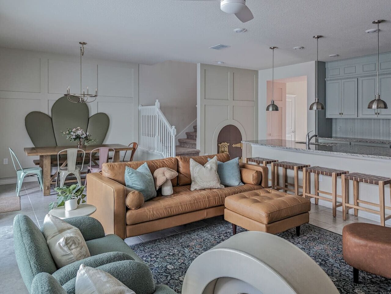 Un salon moderne avec un canapé en cuir marron, des fauteuils verts et une table basse ronde. On y trouve également un coin cuisine avec des meubles en bois et un éclairage suspendu.