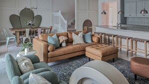 Una sala de estar moderna con un sofá de cuero marrón, sillones verdes y una mesa de centro redonda. Cuenta con una zona de cocina con armarios de madera e iluminación colgante.