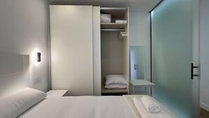 Une chambre d’hôtel moderne, équipée d’un lit, d’une armoire, d’une petite table et d’une salle de bain visible à travers une porte en verre.