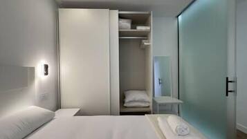 A modern hotel room with a bed, a wardrobe, a small table, and a bathroom visible through a glass door.