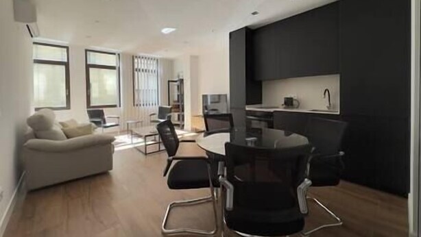 A modern living room with a sofa, a dining table with chairs, a kitchen area with dark cabinets, and large windows providing natural light.