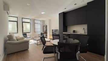 A modern living room with a sofa, a dining table with chairs, a kitchen area with dark cabinets, and large windows providing natural light.
