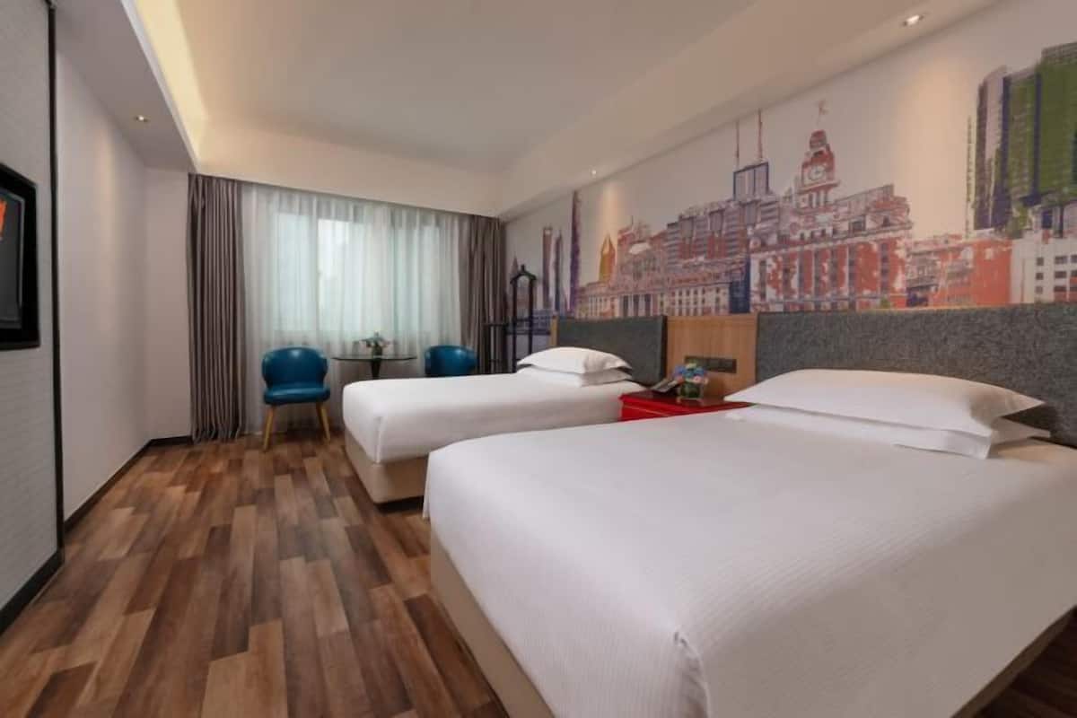 A hotel room with two beds, a cityscape mural, a wooden floor, a blue chair, and a small table.