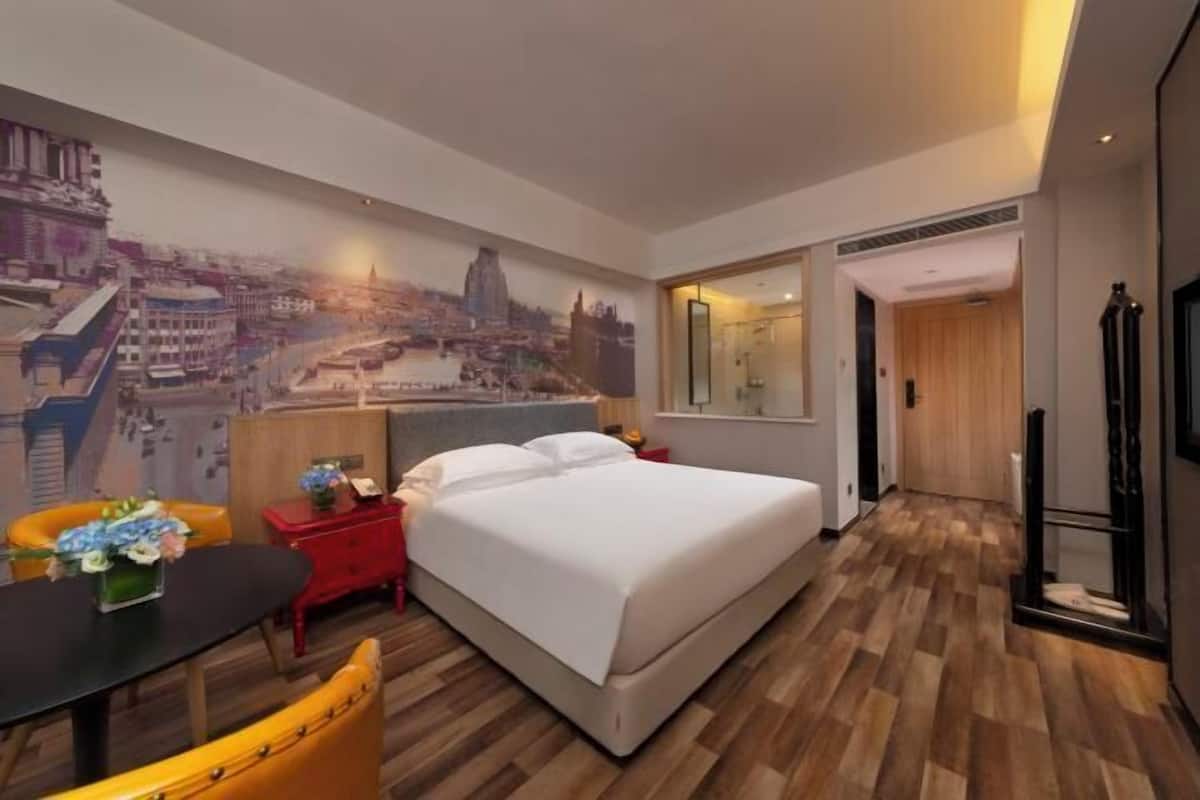 A hotel room with a bed, a desk, a chair, a mirror, and a cityscape mural on the wall.