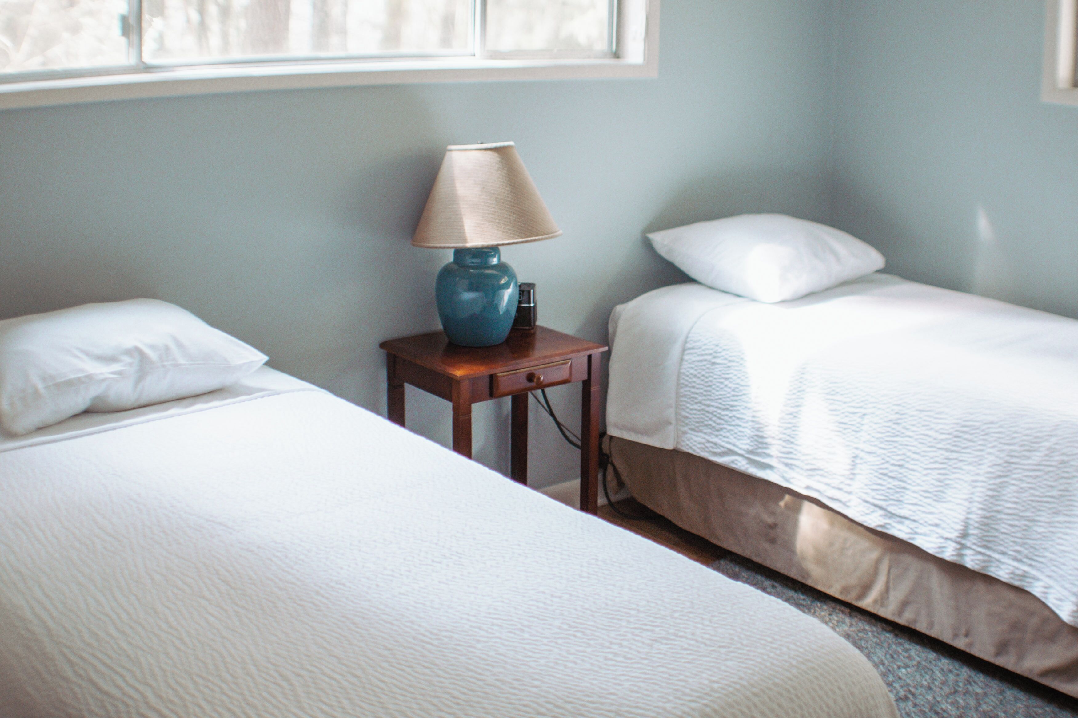 A hotel room with two beds, a nightstand with a lamp, and a window providing natural light.