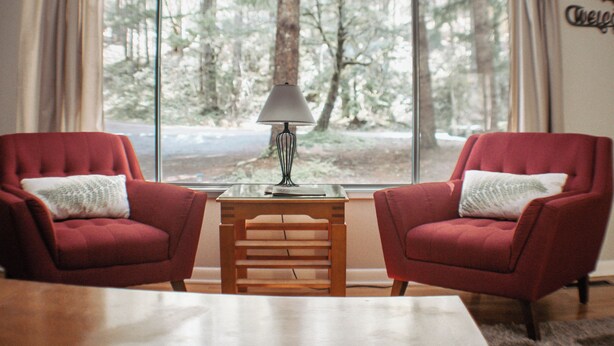 Ein Zimmer mit zwei roten Sesseln, einem Holztisch mit Lampe und einem großen Fenster mit Blick auf einen Wald.