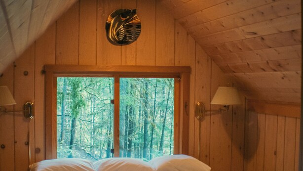 Un dormitorio de cabaña de madera con una cama, dos lámparas fijadas a la pared y un ventanal con vistas a un bosque.