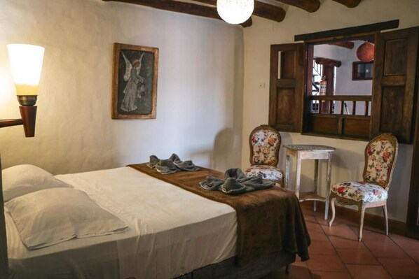 Quarto com cama, duas cadeiras, uma mesinha, abajur e um quadro na parede.
