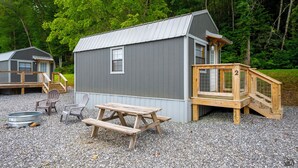 Een kleine houten hut met een terras en picknicktafel, omgeven door grind en bomen.