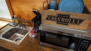 A kitchen with a stainless steel sink, a microwave, and a wooden box labeled "CHEVROLET" containing blue plates and utensils. - 1 Room Cabin- " The Garage" (Robbinsville)