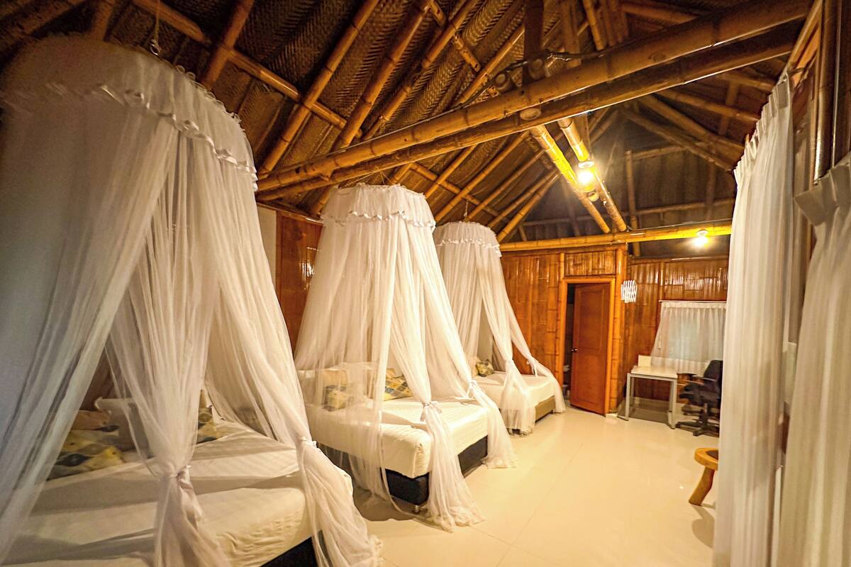 A room with four beds, each covered with white canopy beds. The room has a wooden ceiling and walls, and a small table with a chair.