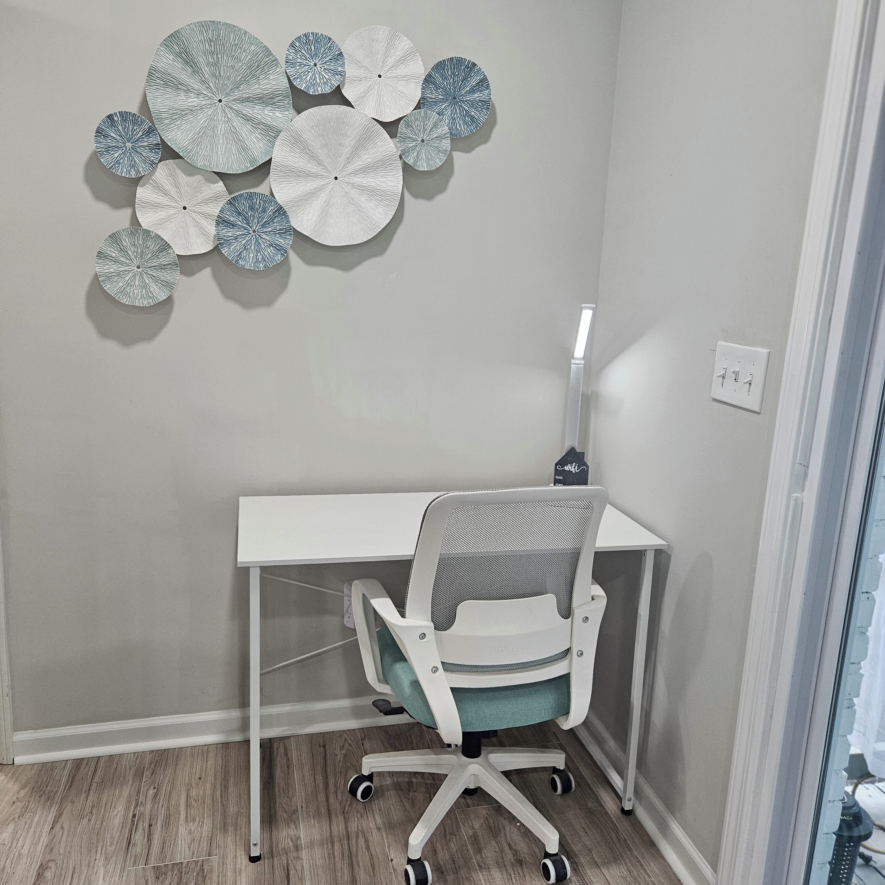 A small office space with a white desk, a mesh office chair, and a wall-mounted light.