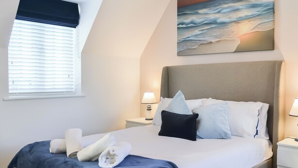 A hotel room with a bed, a nightstand, a lamp, a painting of a beach scene, and a window with blinds. - 3 bedroom accommodation in Filey (Filey)