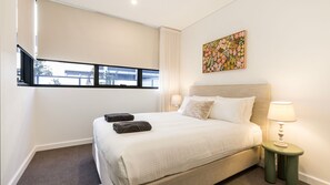 A hotel room with a bed, two pillows, a nightstand with a lamp, a window with blinds, and a floral artwork on the wall. - Iggy's Retreat Huskisson by Latitude South Coast (Huskisson)