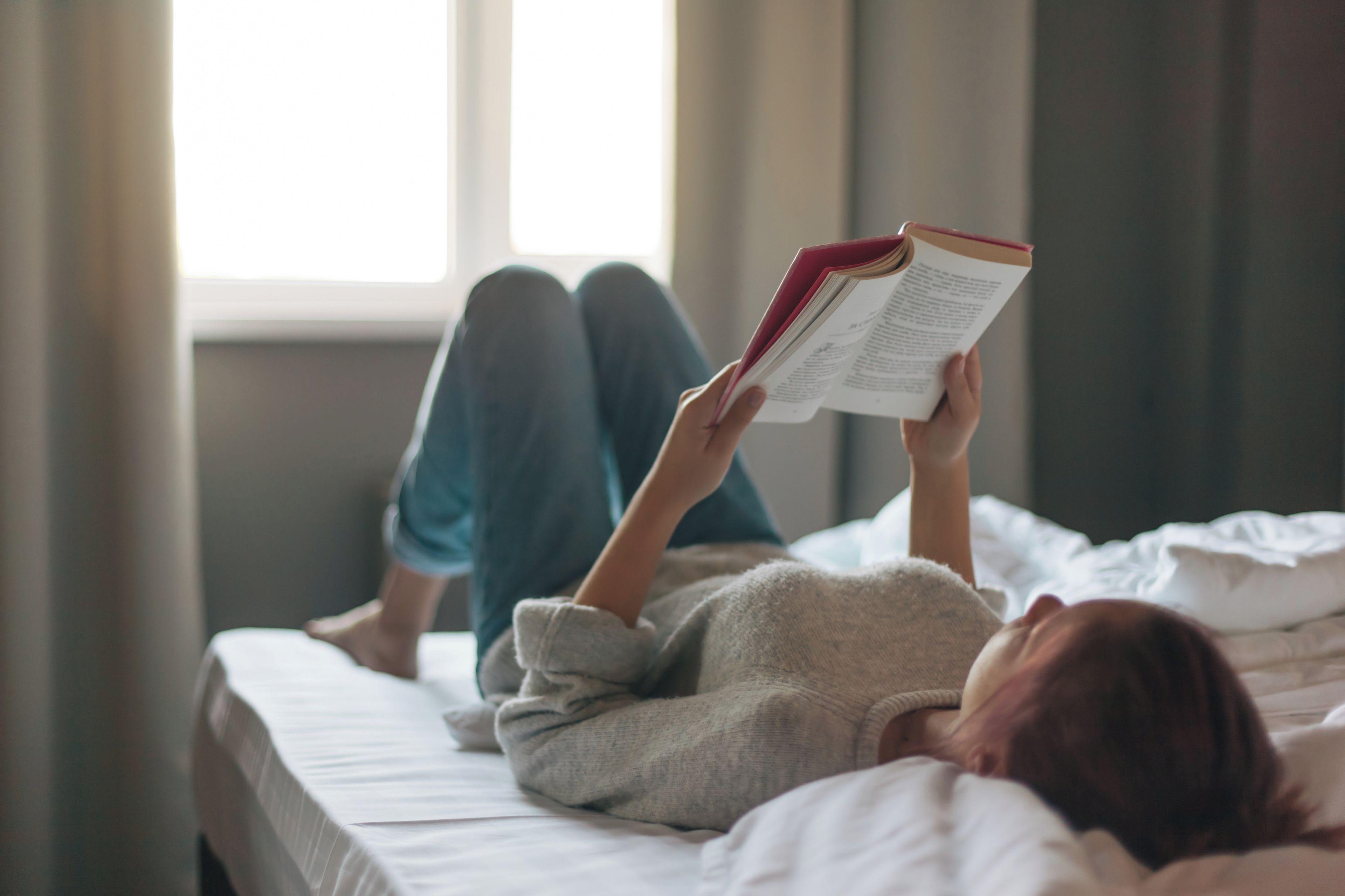 Uma pessoa deitada na cama com um livro, em um quarto com janela e cortinas.