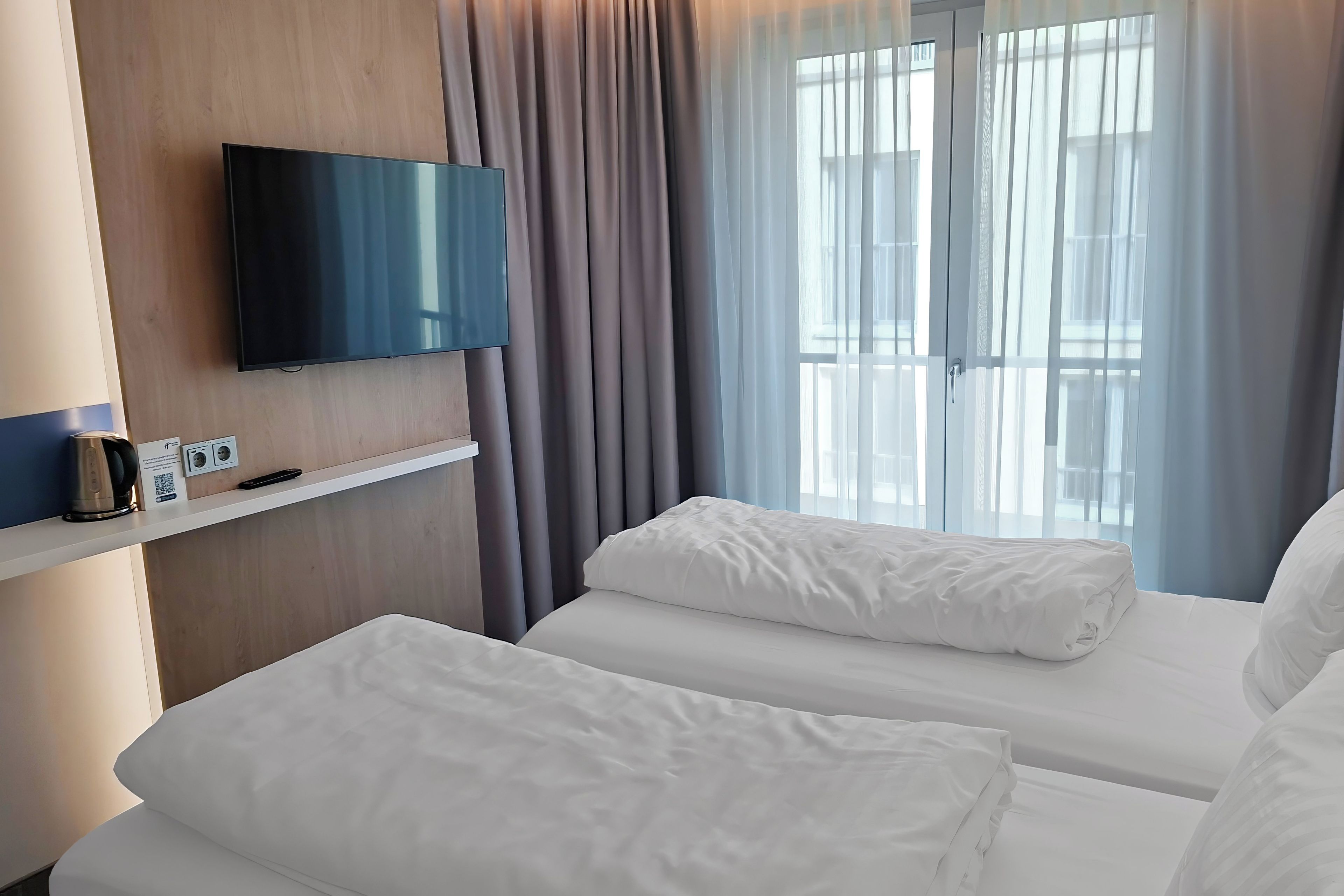 A hotel room with a flat-screen TV mounted on the wall, a bedside table with a kettle and a remote control, two beds with white linens, and a window with sheer curtains.