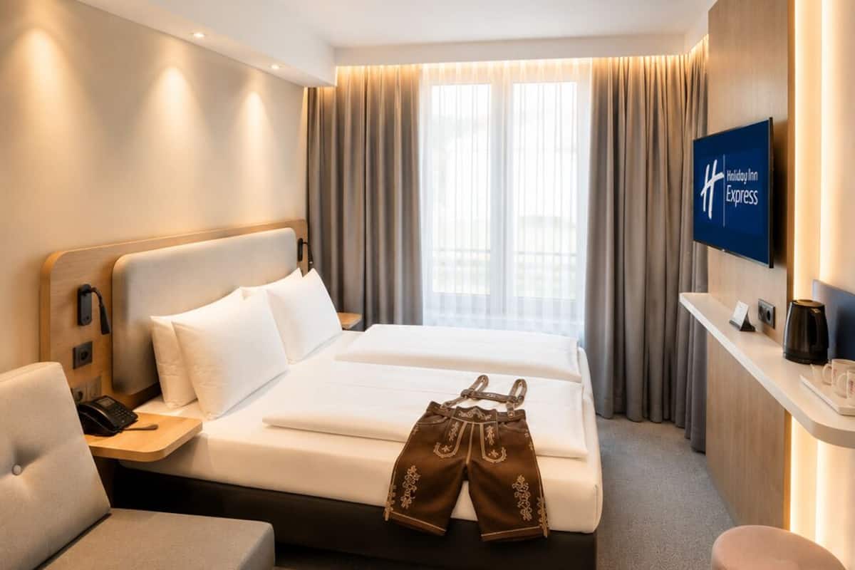 A hotel room with a bed, a desk, a chair, a TV, and a sign on the desk.