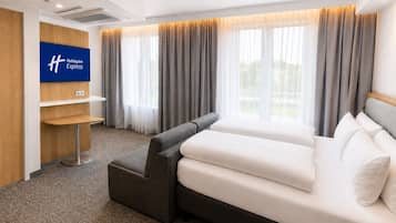 A hotel room with a bed, a desk, a chair, a TV, and large windows with curtains.