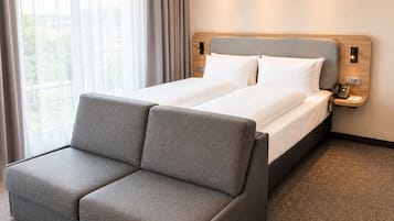 Una habitación de hotel con cama, un sofá gris, una mesita de noche con teléfono y una ventana con cortinas.