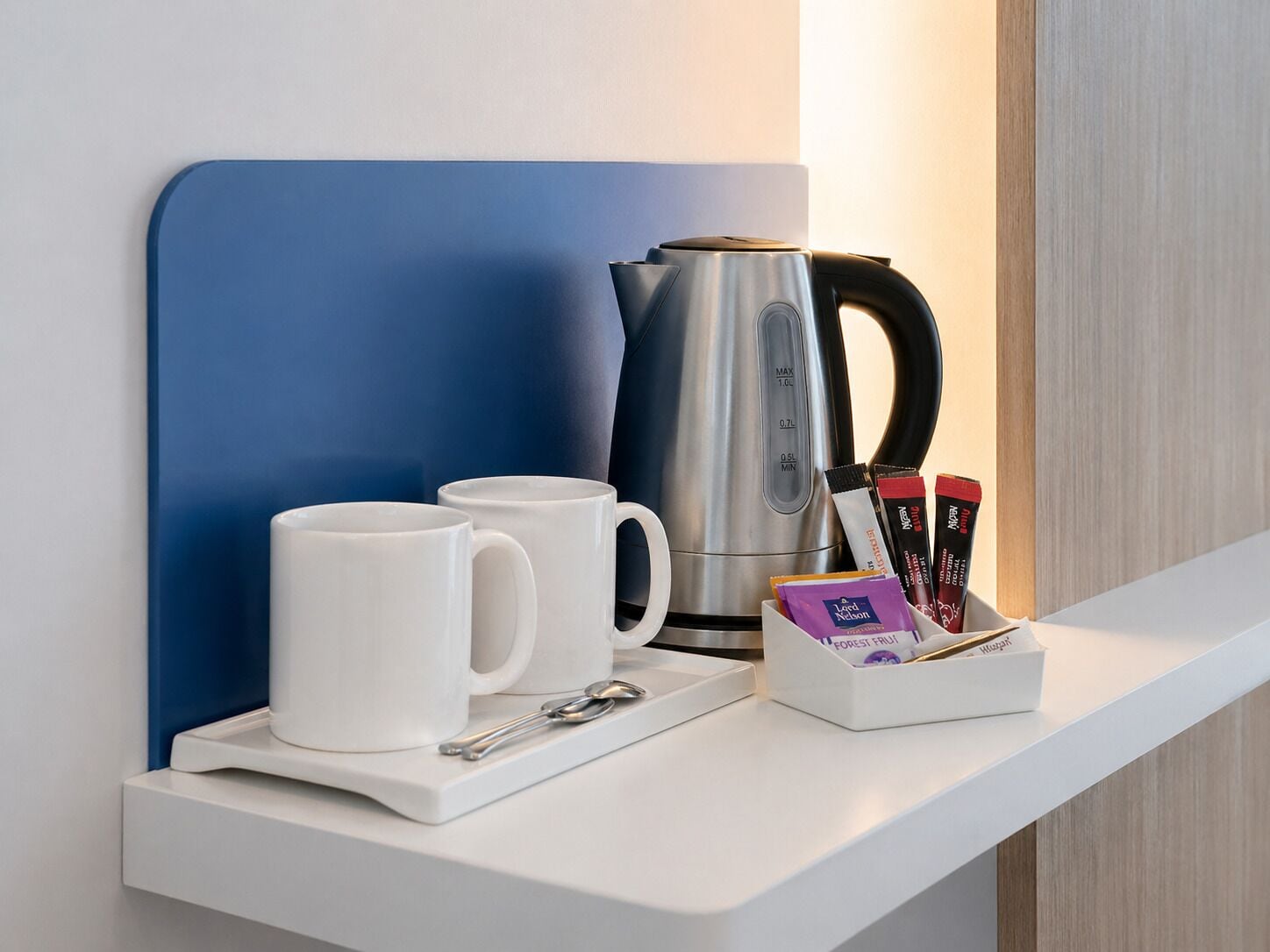 A white countertop with a blue wall, a silver kettle, two white mugs, a spoon, and a box of tea bags.