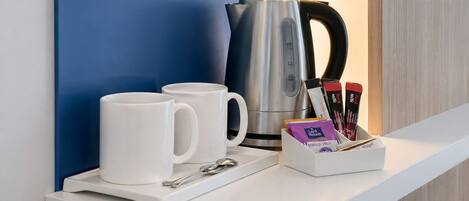 A white countertop with a blue wall, a silver kettle, two white mugs, a spoon, and a box of tea bags.