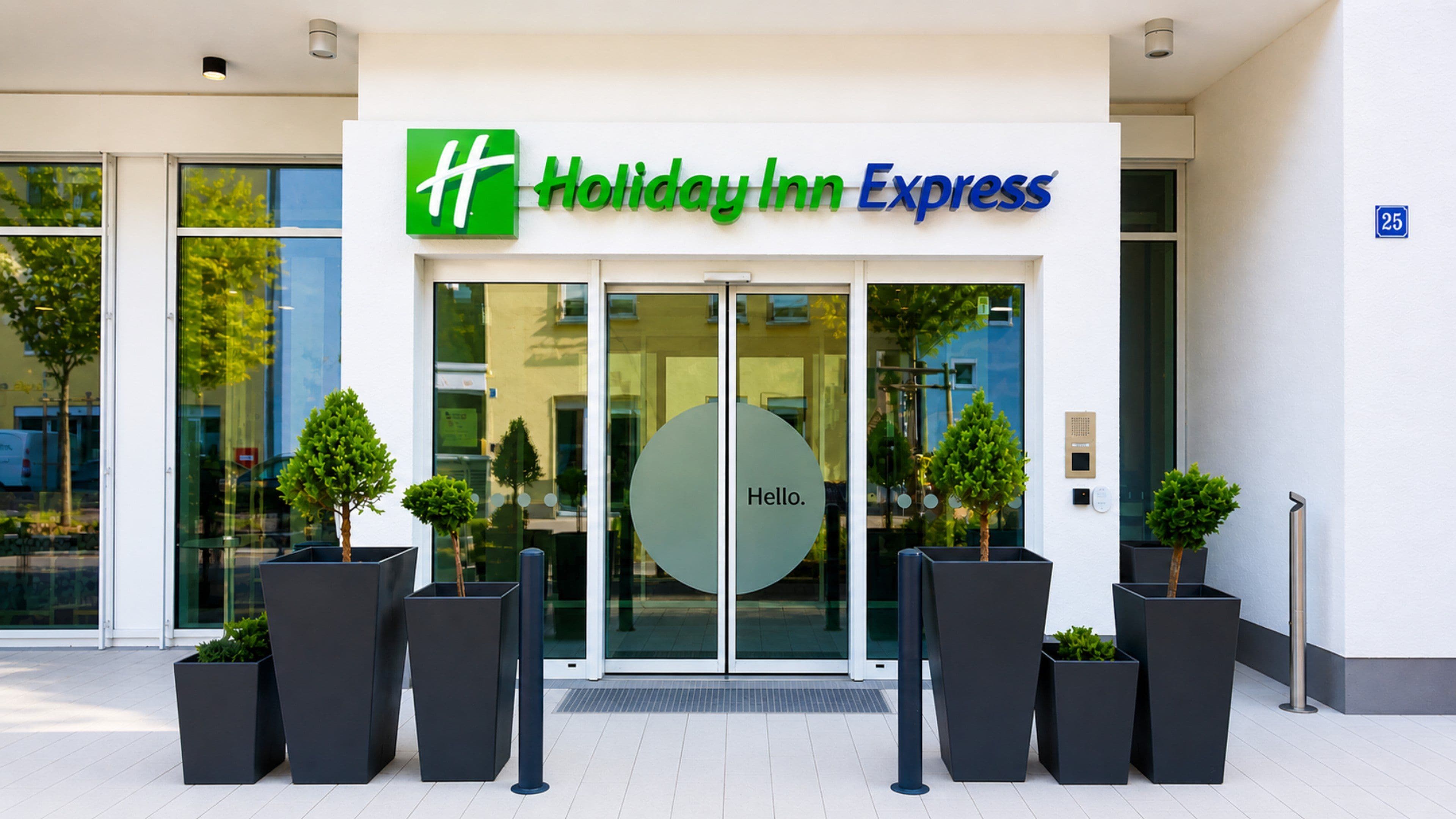 Edificio de Holiday Inn Express con entrada de vidrio, un letrero que dice 'Hello' y plantas en macetas al frente.