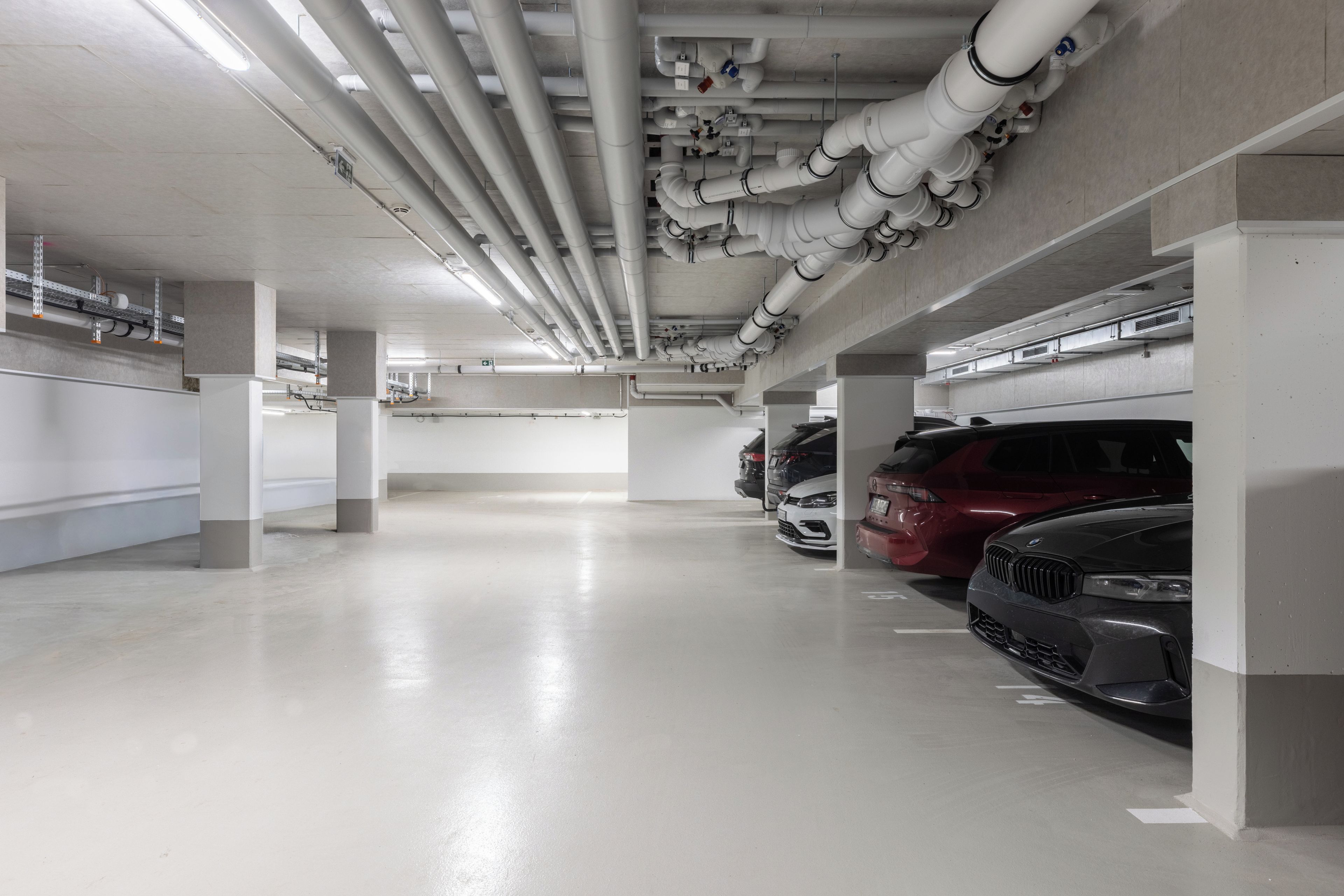 Un estacionamiento de varios niveles con pilares de hormigón, tuberías elevadas y varios automóviles estacionados.