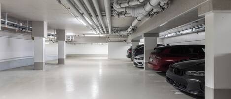 Un estacionamiento de varios niveles con pilares de hormigón, tuberías elevadas y varios automóviles estacionados.