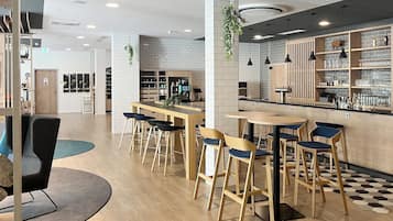 Un interior de cafetería moderna con mesas y sillas de madera, una zona de bar con estanterías llenas de vasos, y un suelo con estampado de cuadros.