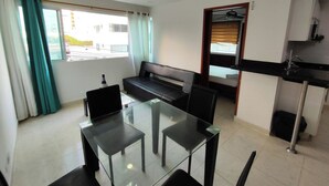 A compact living space with a glass dining table, black chairs, a black sofa, and a kitchenette area. - EM3-1 APARTAMENTO 1 HAB ZONA RESIDENCIAL (Cartagena)