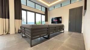 Een moderne woonkamer met een grote bank, een flatscreen televisie en uitzicht op het buitenleven door grote ramen.