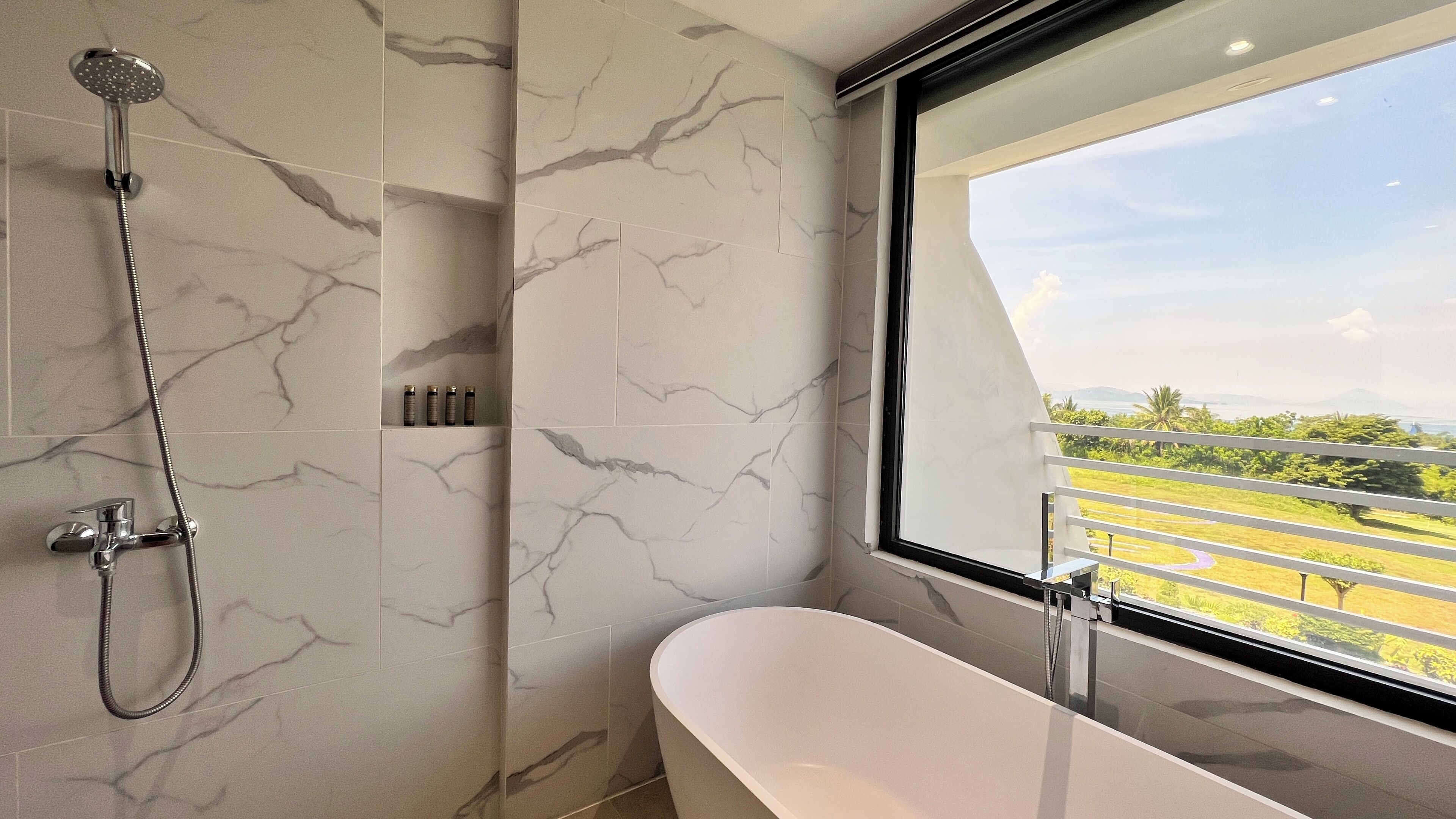 A modern bathroom with a freestanding bathtub, a showerhead, and a window offering a view of a landscaped area.