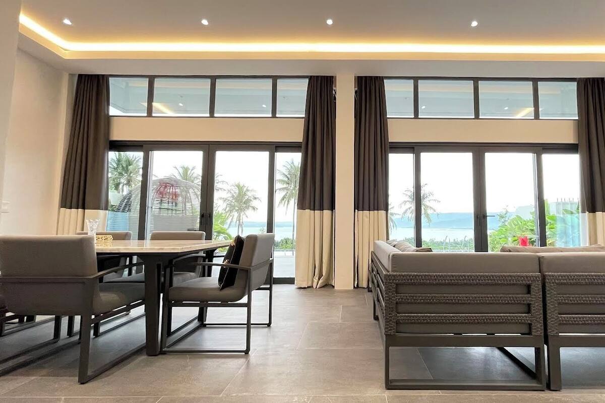 Una habitación de hotel moderna con zona de comedor, sofá y grandes ventanales con vista al mar.
