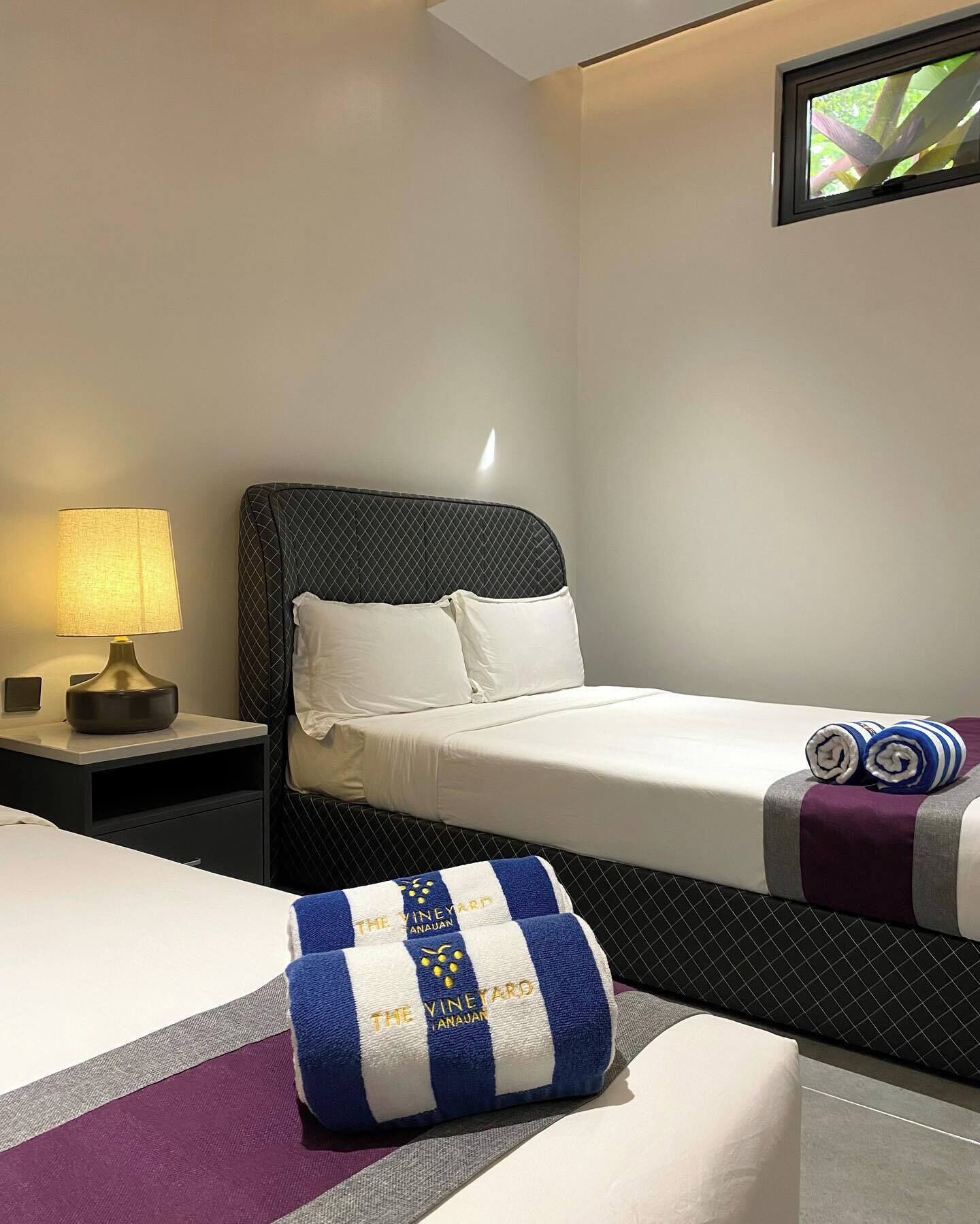 A hotel room with a bed, a nightstand with a lamp, and a window with a view of greenery.